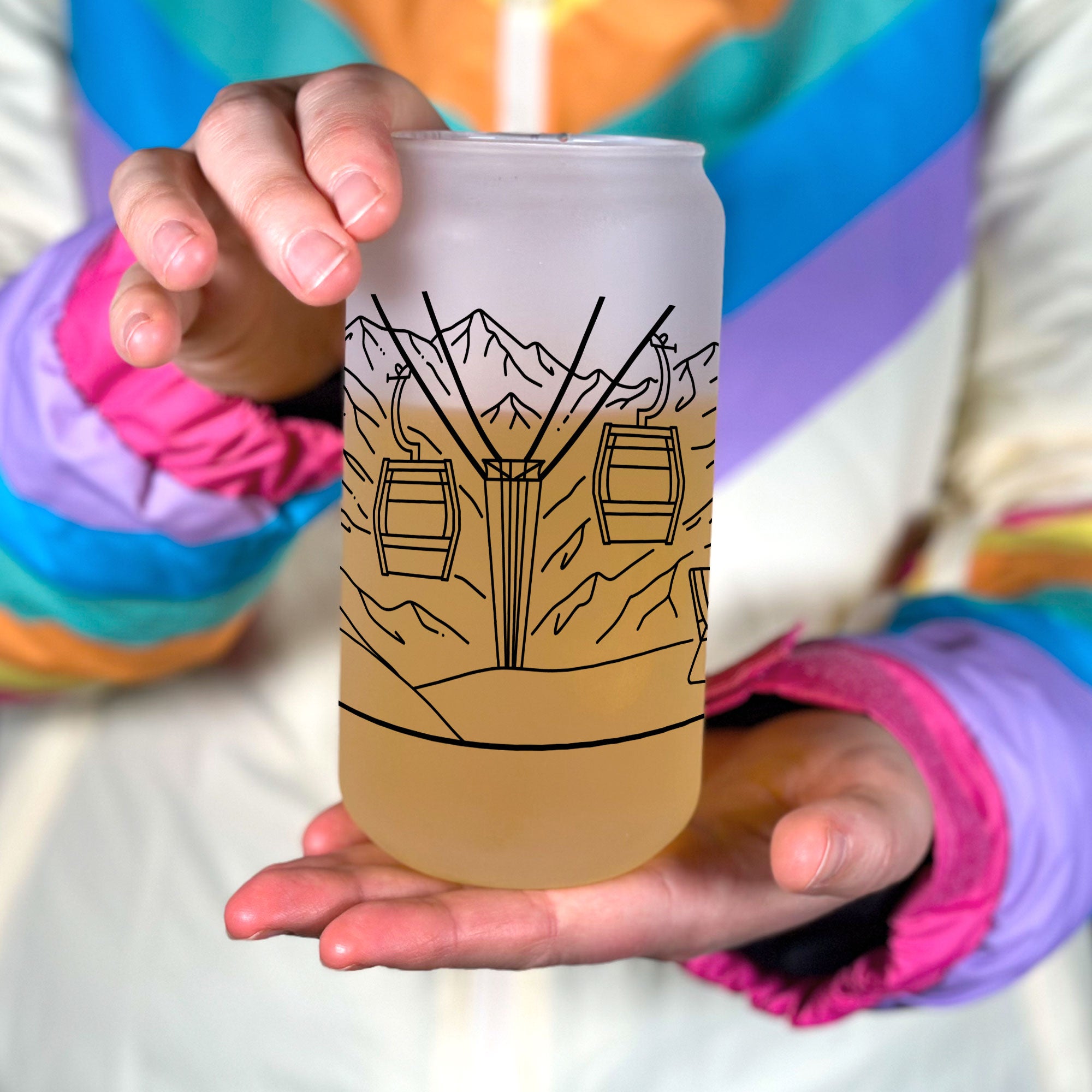 Hand holiding frosted glass tumbler. Shows middle ski couple line art design of ski chair lift.