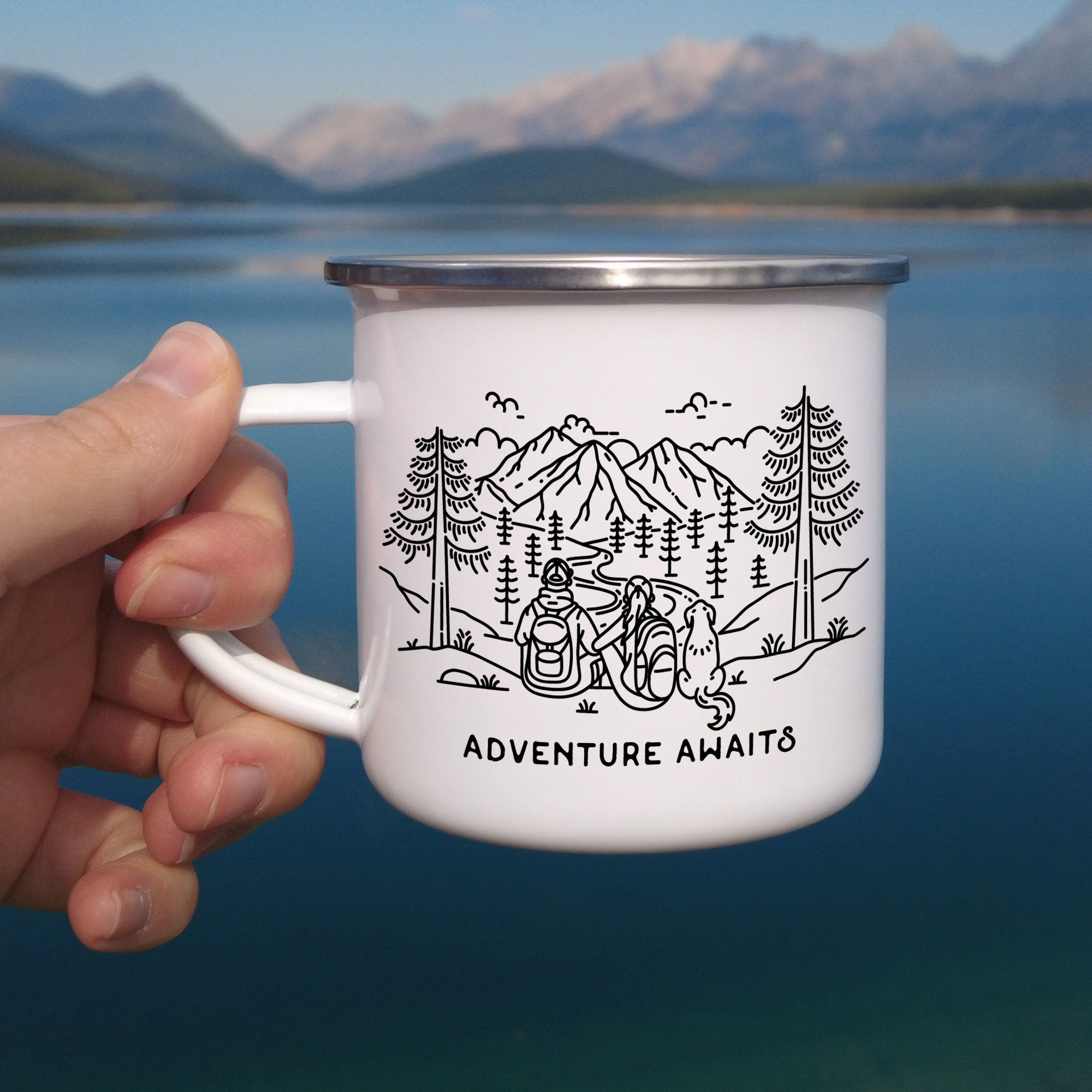 Close-up of hand holding single silver rimmed enamel mug. The mug features line artwork of a mountain hiking couple with their floppy eared dog (looks like a gold retriever). The front text reads Adventure Awaits.