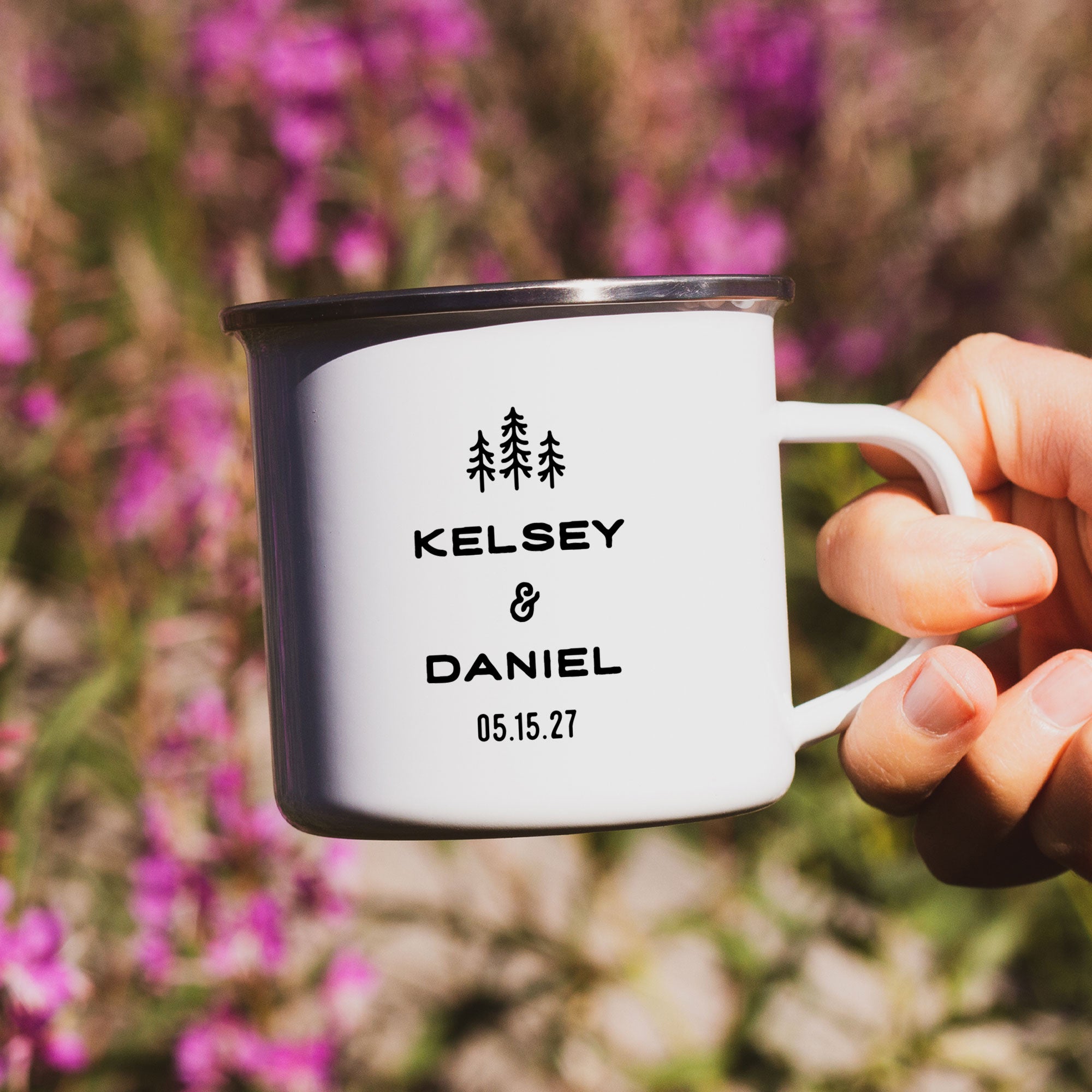 Close-up of hand holding single silver rimmed enamel mug. Photo references the back (or right side) of the enamel mug design. The back has 3 Line Art Trees and the text 'Kelsey & Daniel 05.15.27'