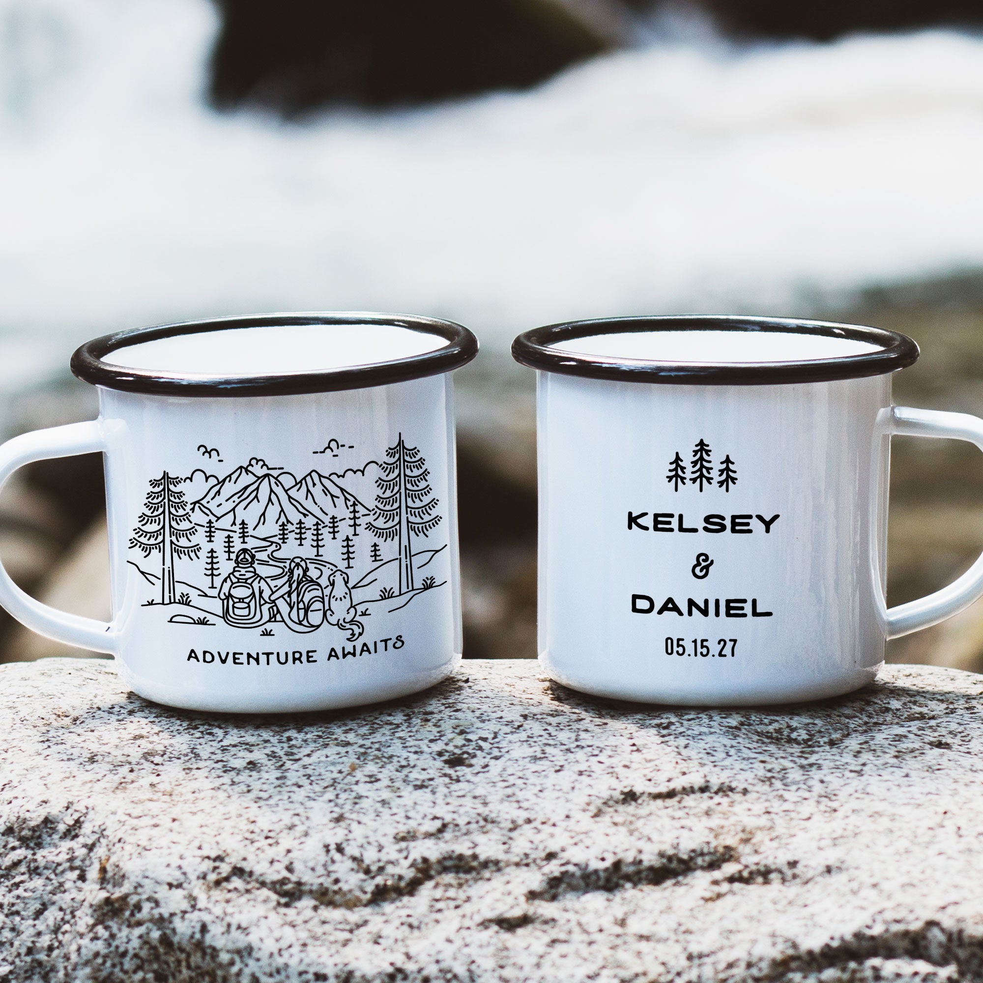 Two black rimmed enamel mugs on river rock. Mug design features line artwork of mountain hiking couple and dog (droppy earred dog similar to a golden retriever). Front text says 'Adventure Awaits'. Back design with 3 Line Art Trees and text 'Kelsey & Daniel 05.15.27'