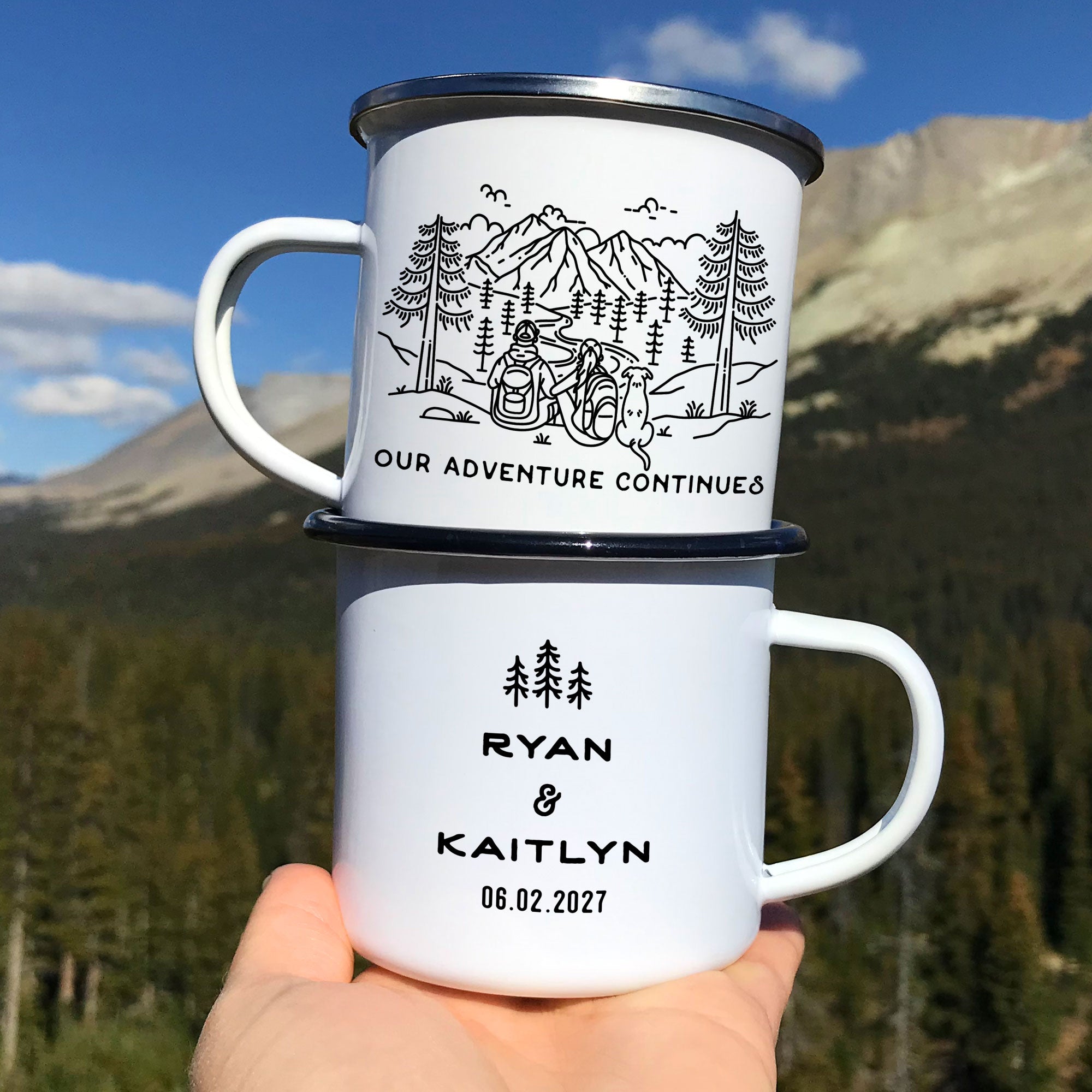 Hand holding two stacked black & silver rimmed enamel mugs. Mug design features line artwork of mountain hiking couple and dog (floppy earred dog). Front text says 'Our Adventure Continues'. BaTck design with 3 Line Art rees and text 'Ryan & Kaitlyn 06.02.2027'