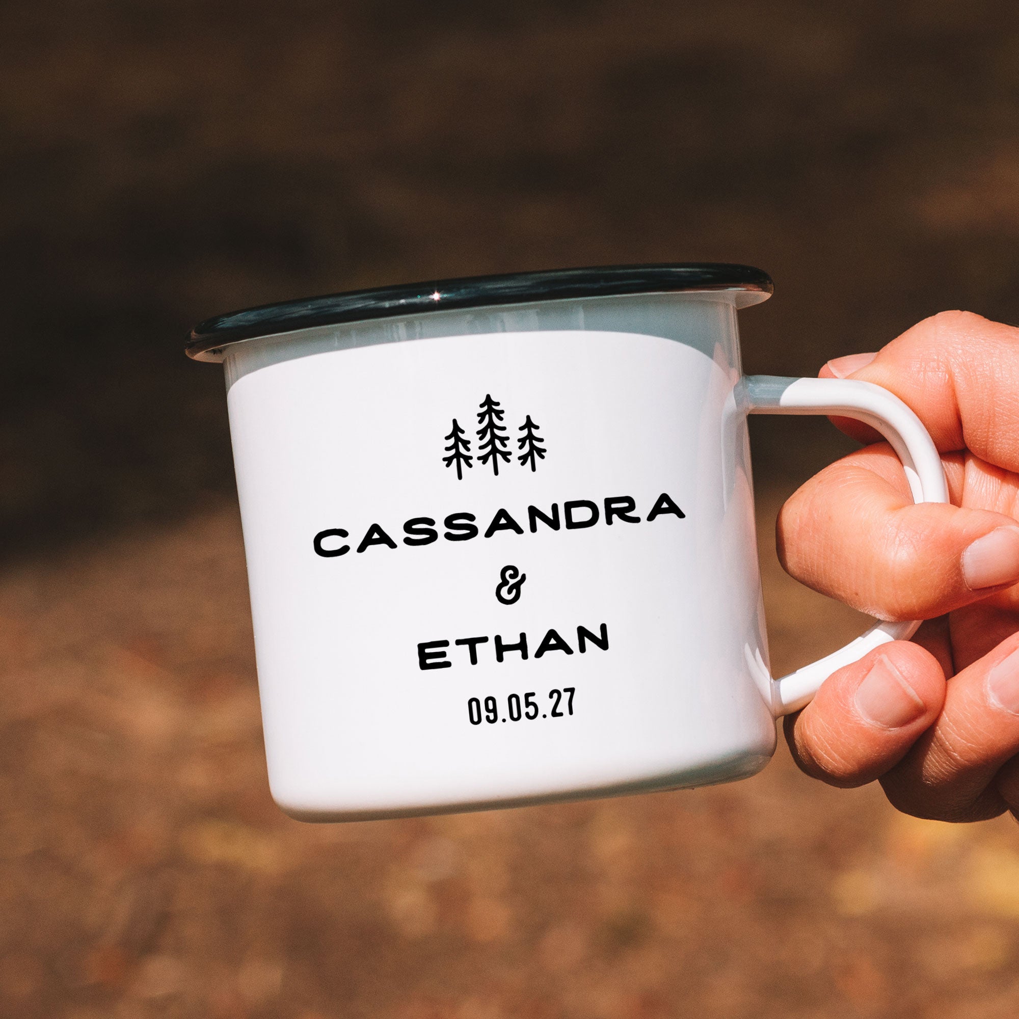 Close-up of hand holding single black rimmed enamel mug. Photo references the back (or right side) of the enamel mug design. The back has 3 Line Art Trees and the text 'Cassandra & Ethan 09.05.27'