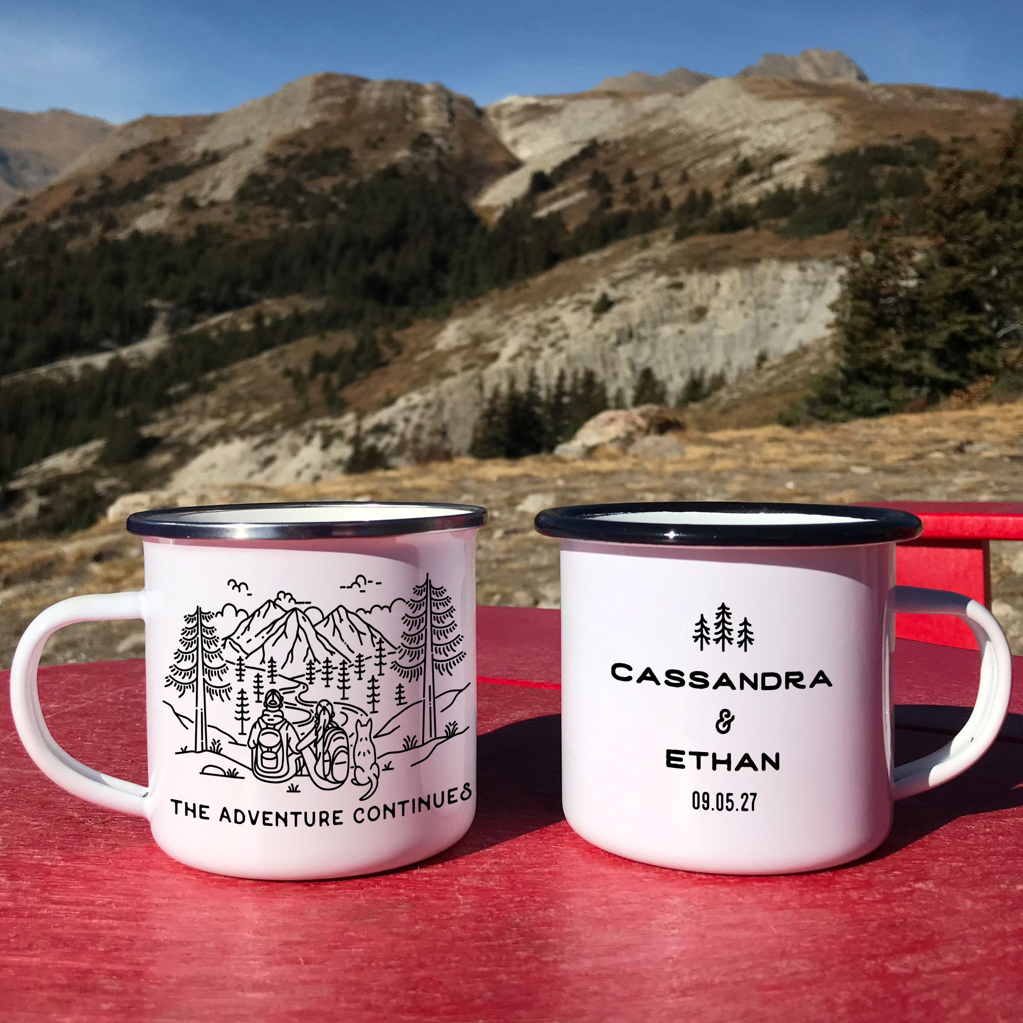 Two black & silver rimmed enamel mugs placed on red chair. Mug design features line artwork of mountain hiking couple and pointy eared dog. Front text says 'The Adventure Continues'. Back design with 3 Line Art rees and text 'Cassandra & Ethan 09.05.27'