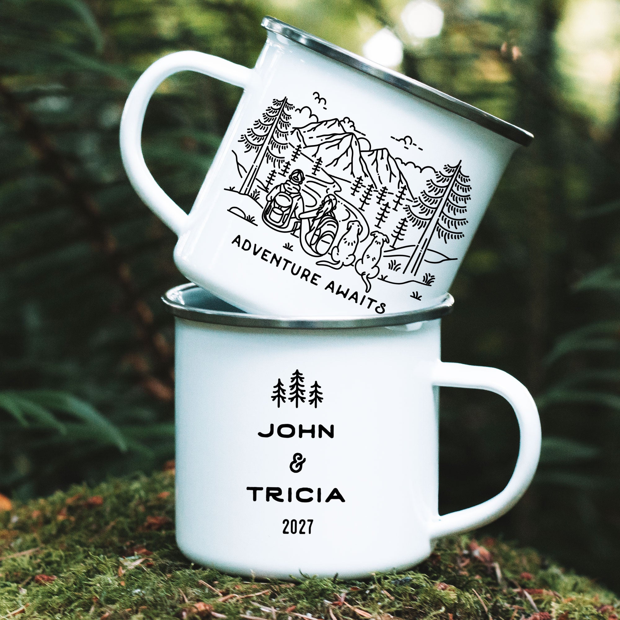 Stacked silver rimmed enamel mugs on mossy rock. Features original hand drawn line artwork of mountain hiking couple & two floppy earred dogs. Text on front of mug shows 'Adventure Awaits'. Design on back of mug is of three line art trees and the text 'John & Tricia 2027'