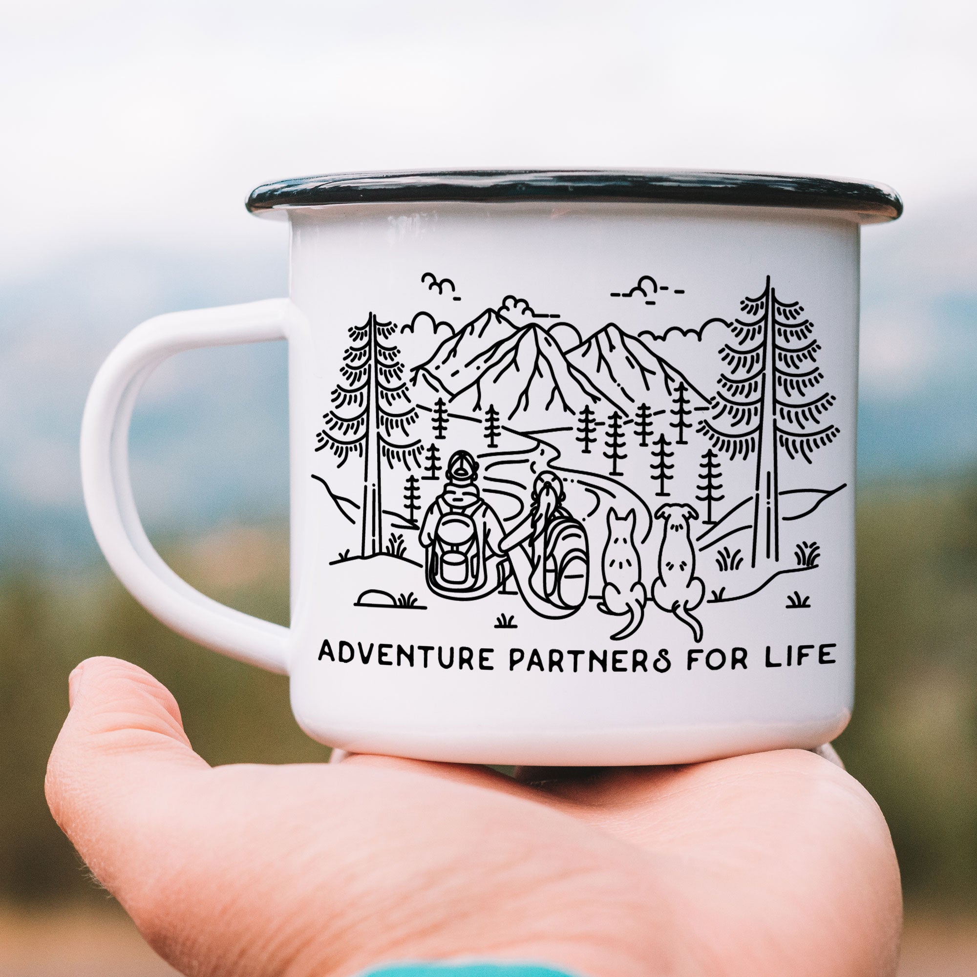 Hand holding single black rimmed enamel mug. The mug features line artwork of a mountain hiking couple with their two dogs (1 pointy ear & 1 floppy ear). The front text reads Adventure Partners for Life.
