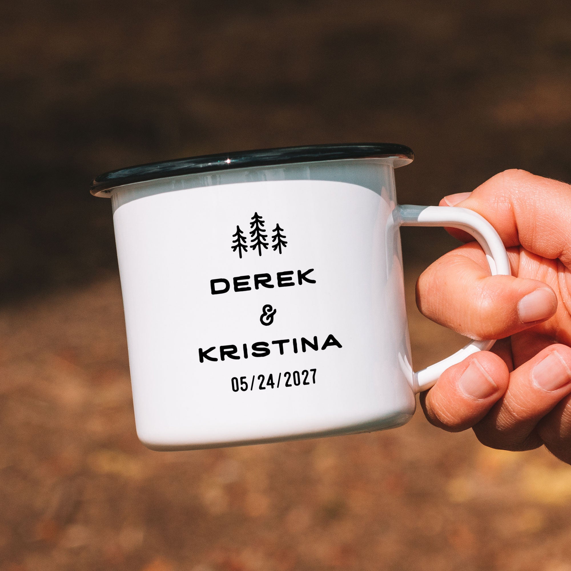 Hand holding single black rimmed enamel mug. Photo references the back (or right side) of the enamel mug design. The back has 3 Line Art Trees and the text 'Derek & Kristina 05/24/2027.'
