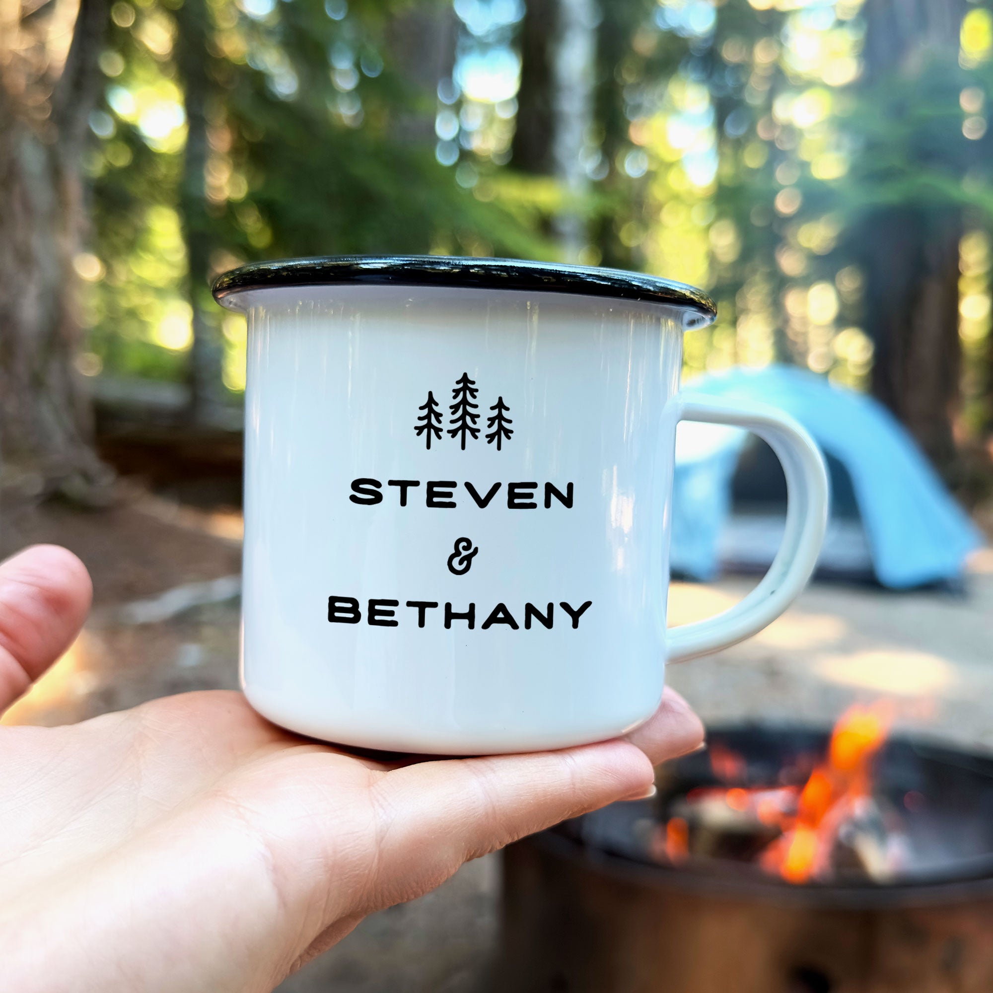 Hand holding single black rimmed enamel mug. Photo references the back (or right side) of the enamel mug design. The back has 3 Line Art Trees and the text 'Steven & Bethany.'
