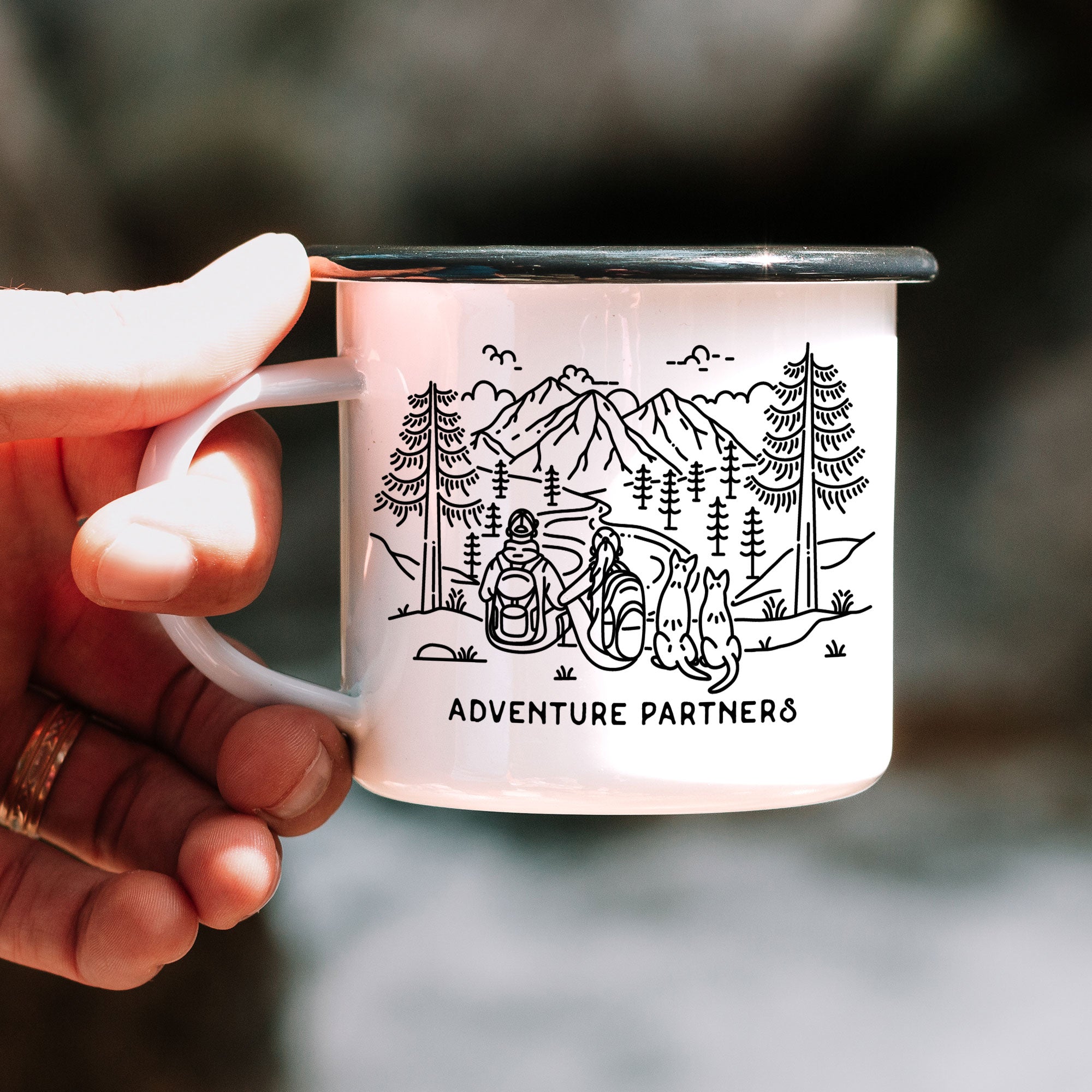 Hand holding single black rimmed enamel mug. The mug features line artwork of a mountain hiking couple with their two pointy eared dogs. The front text reads Adventure Partner.
