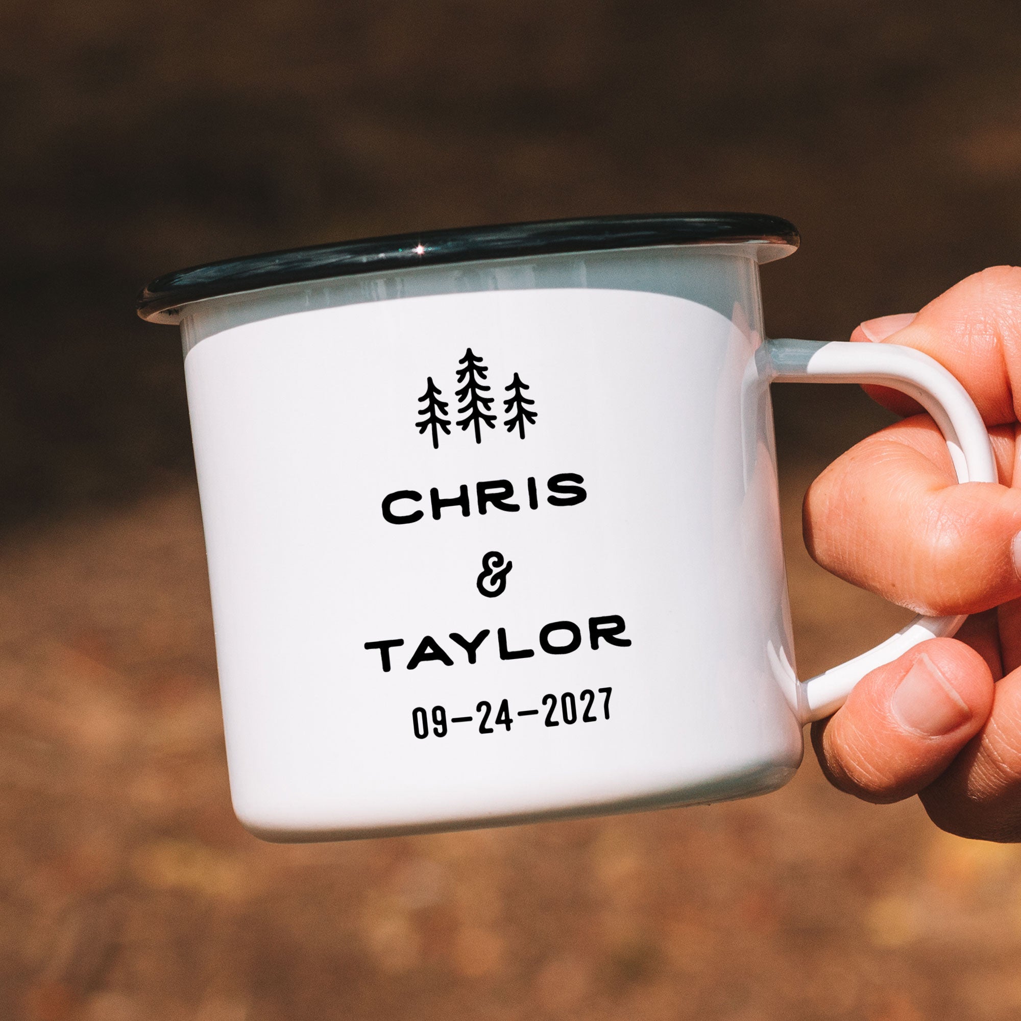 Close-up of hand holding single black rimmed enamel mug. Photo references the back (or right side) of the enamel mug design. The back has 3 Line Art Trees and the text 'Chris & Taylor 09-24-2027.'