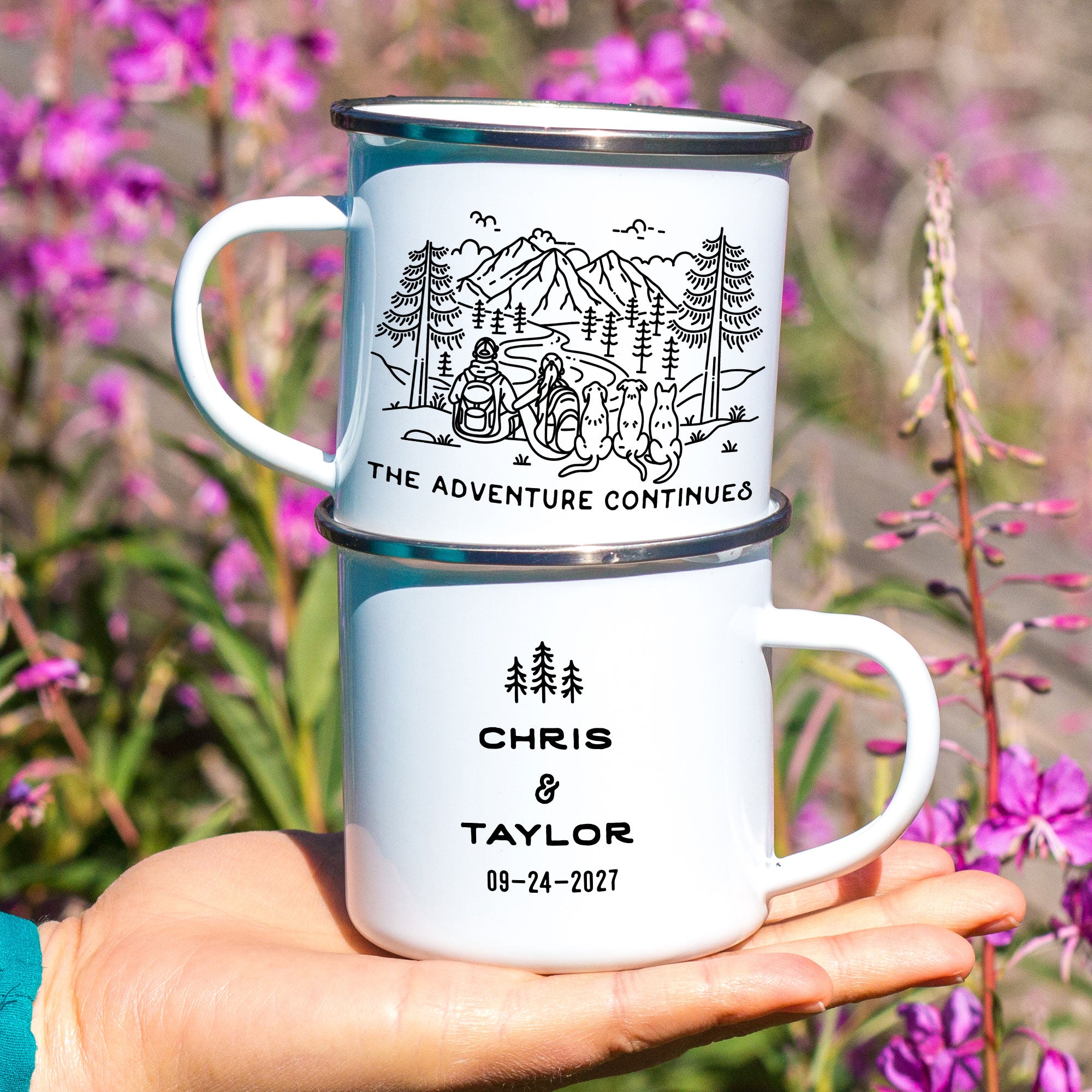 Close-up of hand holding stacked silver rimmed enamel mugs. The mugs feature line artwork of a mountain hiking couple with their 3 dogs. The front text reads 'The Adventure Continues,' and the back has 3 Line Art Trees with the text 'Chris & Taylor 09-24-2027.'