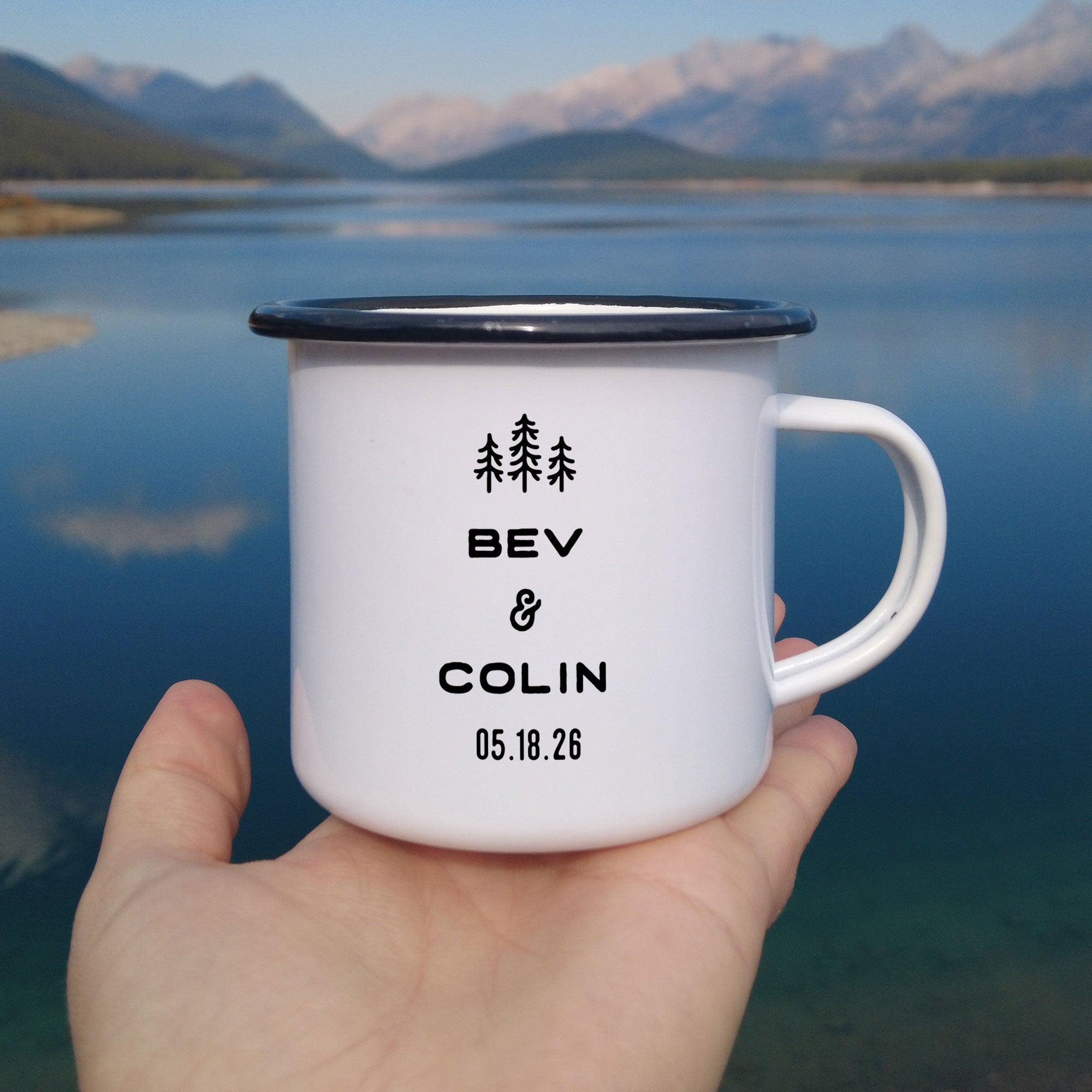 Close-up of hand holding single black rimmed enamel mug with beautiful mountain lake in background. Photo references the back (or right side) of the enamel mug design. The back has 3 Line Art Trees and the text 'Bev & Colin, 05.18.26'