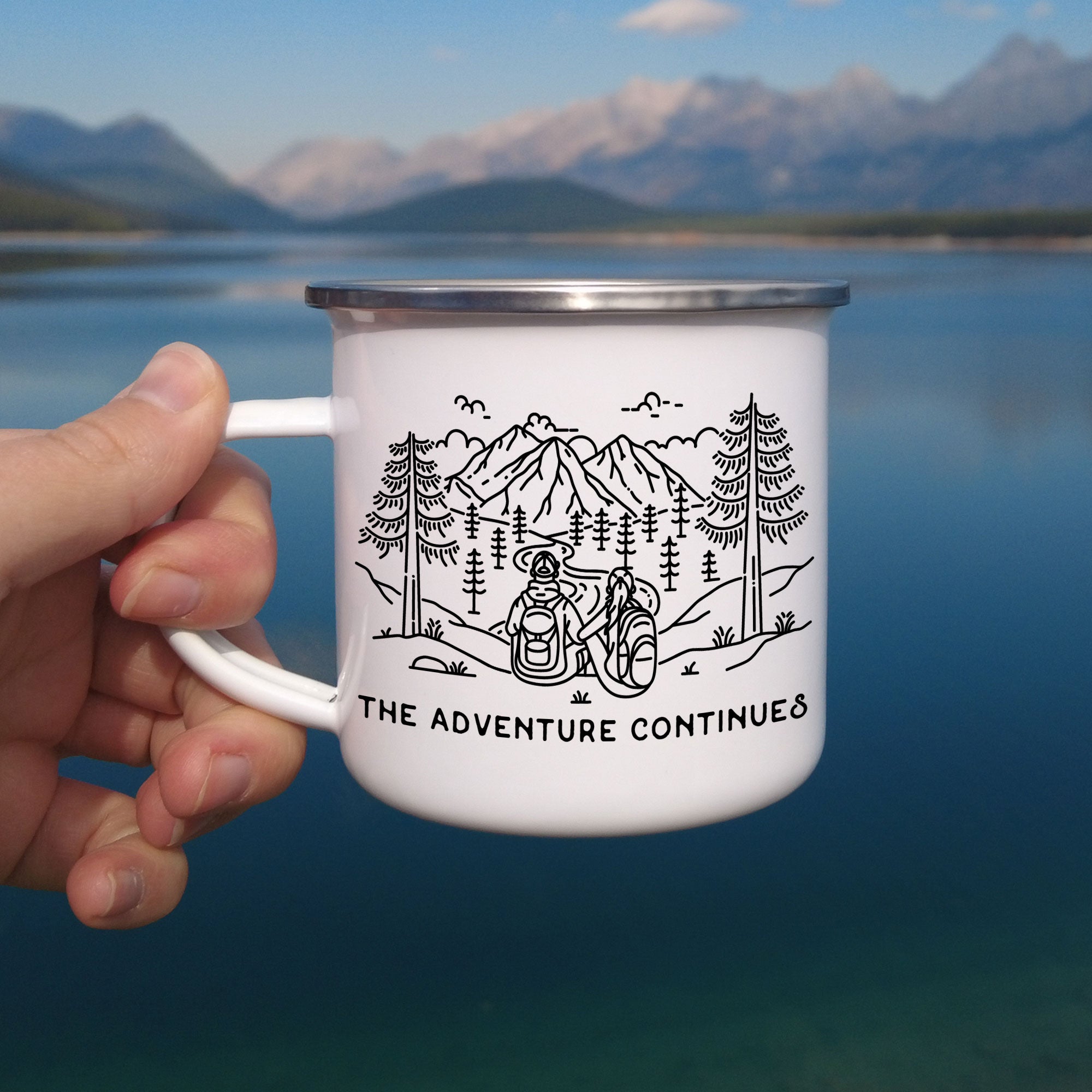 Hand holding one silver rimmed enamel mug. The mug features line artwork of a mountain hiking couple. The front text reads The Adventure Continues.