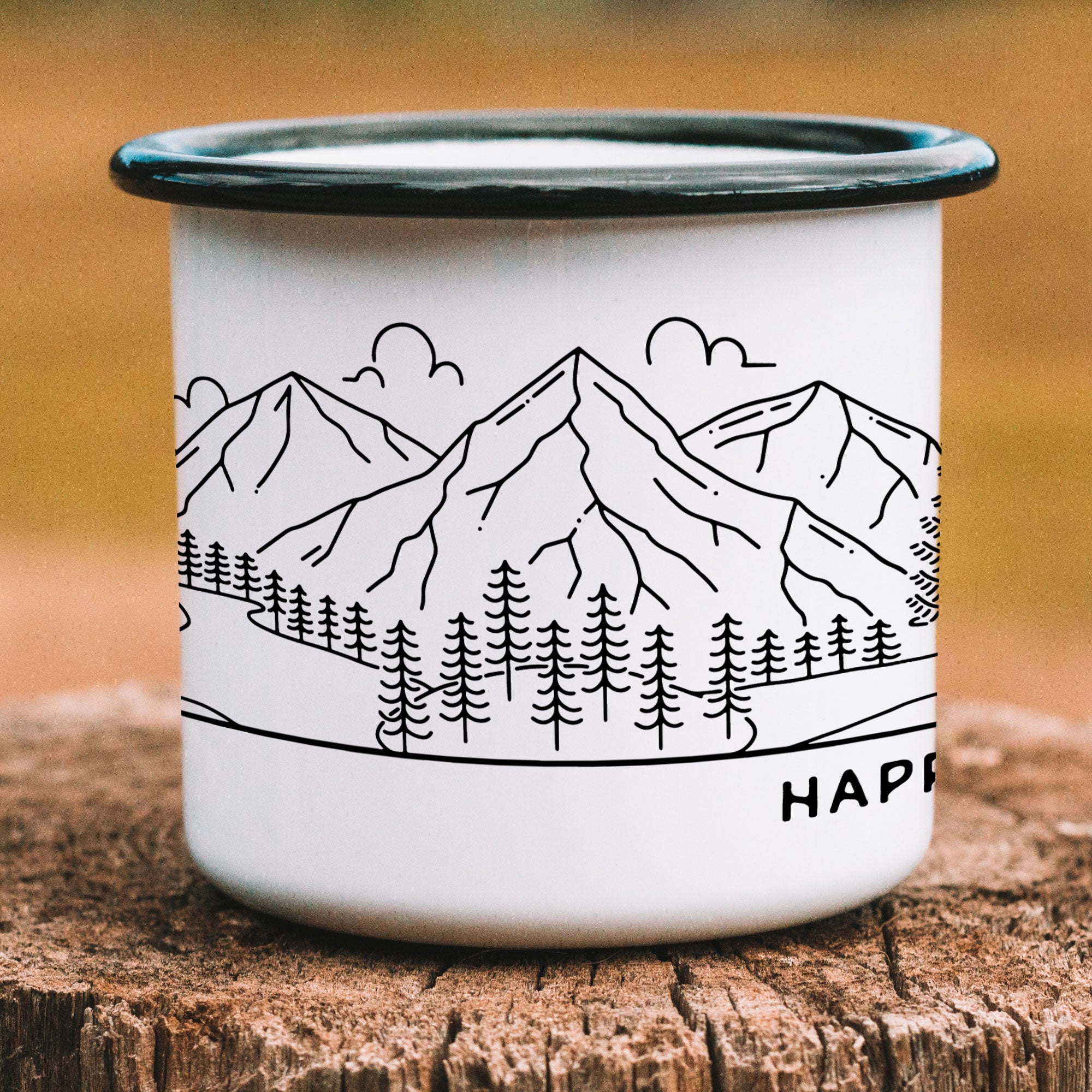 Close-up of black-rimmed enamel mug on wood post. Details center of bride & groom mountain wedding design. Shows mountain range with clouds, lake with trees, and island with more trees.