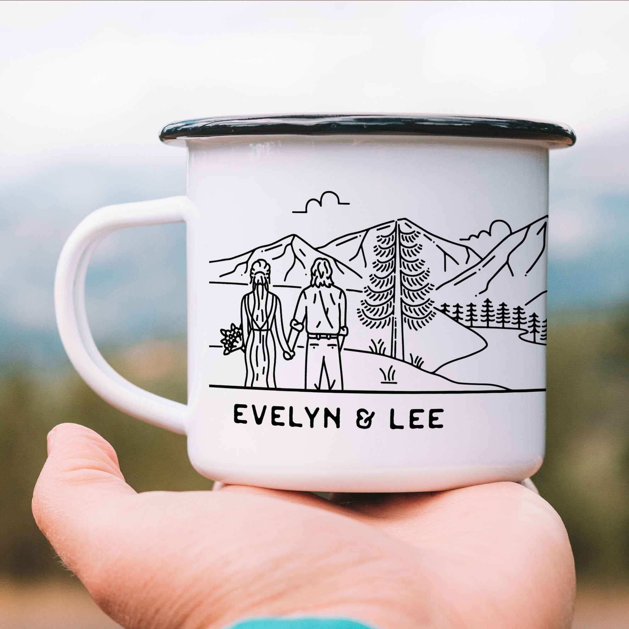 Close-up of hand holding black-rimmed white enamel mug featuring a bride and groom illustration by a mountain lake. The bride has an updo with a veil and carries a floral bouquet. The groom has long hair and wears a dress shirt with rolled-up sleeves.