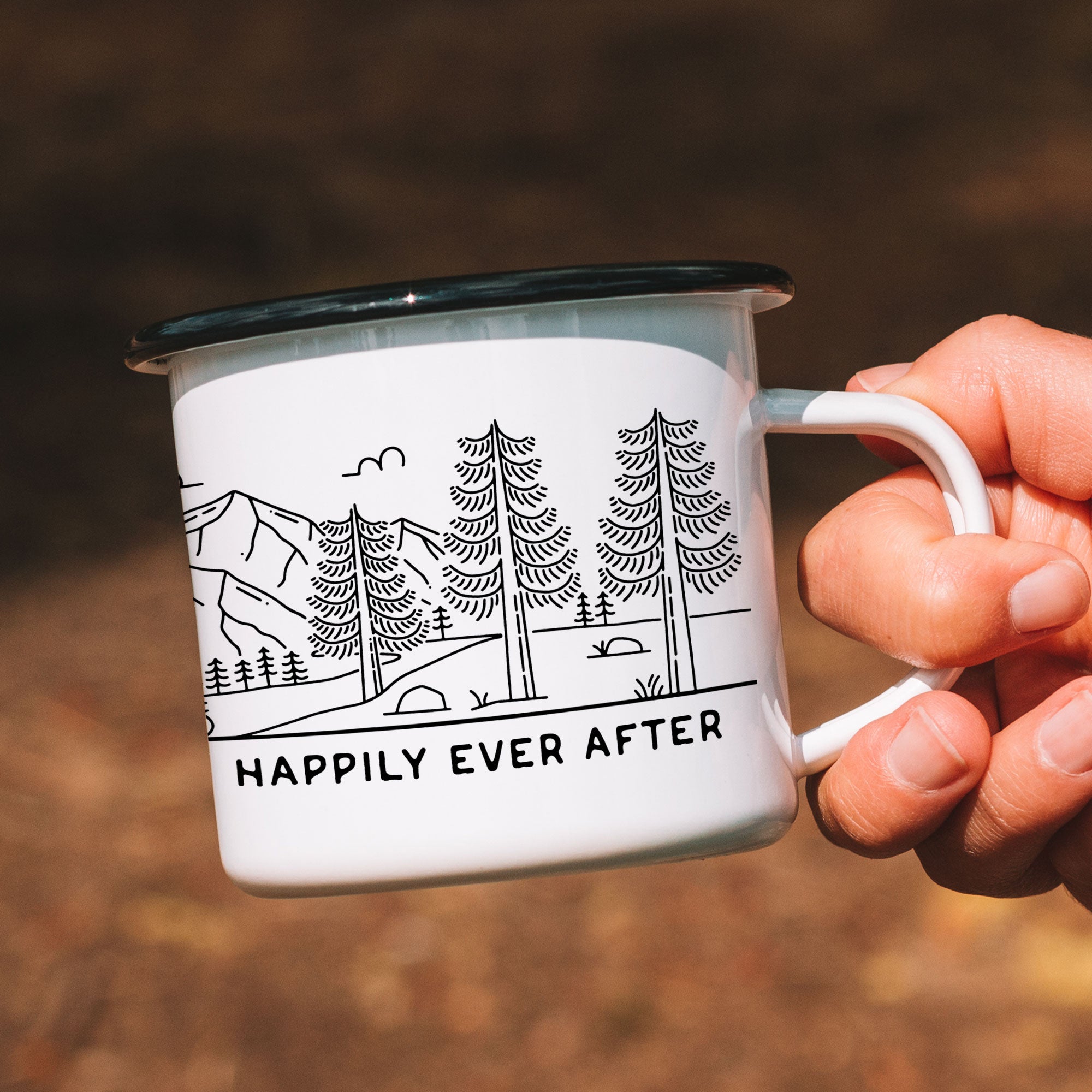 Close-up of black-rimmed white enamel mug with a mountain bride & groom design. Mug shows right side of design which features rocks, grass, and trees.