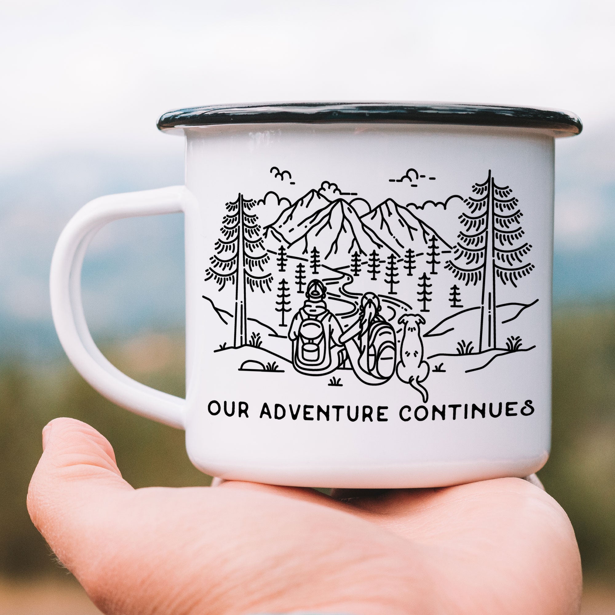 Hand holding single black rimmed enamel mug. The mug features line artwork of a mountain hiking couple with their floppy eared dog. The front text reads Our Adventure Continues.