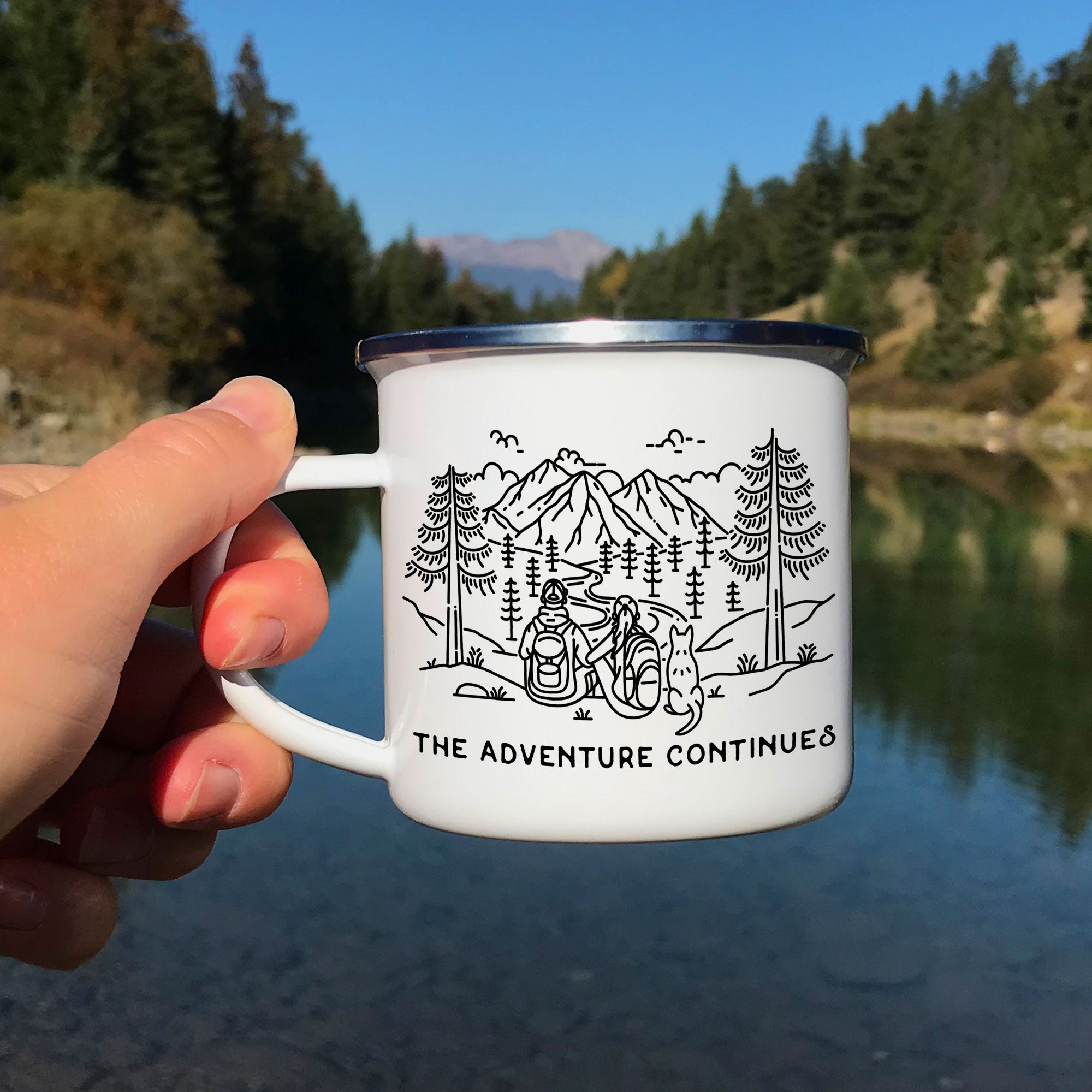 Hand holding single silver rimmed enamel mug. The mug features line artwork of a mountain hiking couple with their pointy eared dog. The front text reads The Adventure Continues.