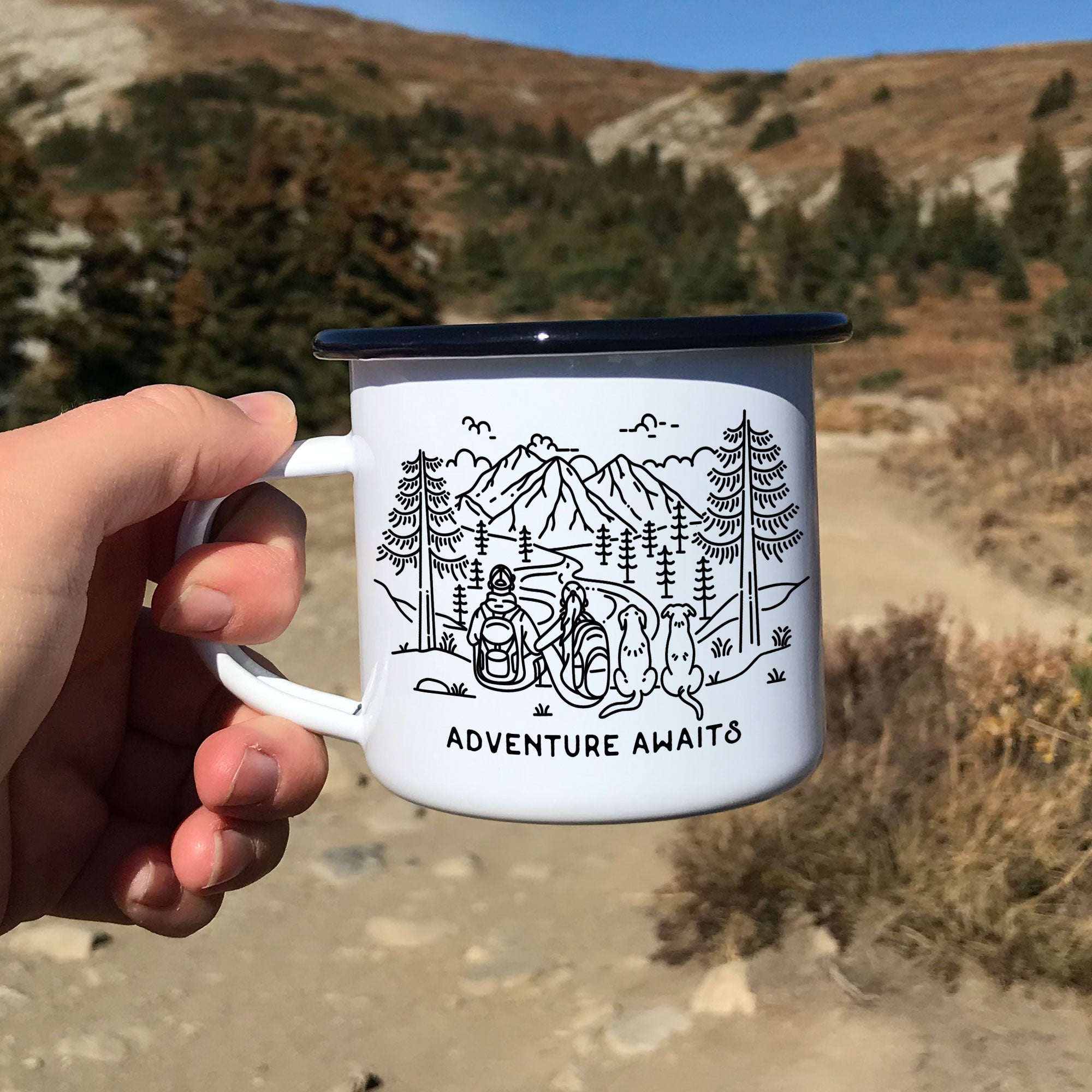 Hand holding single black rimmed enamel mug. The mug features line artwork of a mountain hiking couple with their two floppy eared dogs. The front text reads Adventure Awaits.