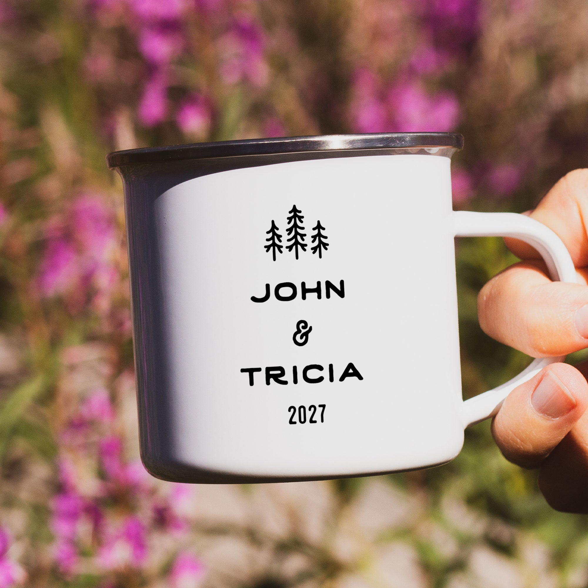 Close-up of hand holding single silver rimmed enamel mug. Photo references the back (or right side) of the enamel mug design. The back has 3 Line Art Trees and the text 'John & Tricia 2027'