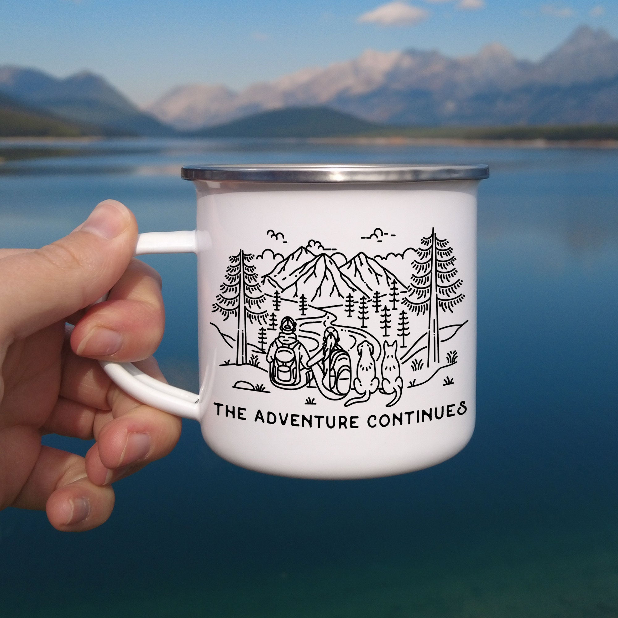 Hand holding single silver rimmed enamel mug. The mug features line artwork of a mountain hiking couple with their two dogs (1 floppy eared & 1 pointy eared). The front text reads The Adventure Continues.