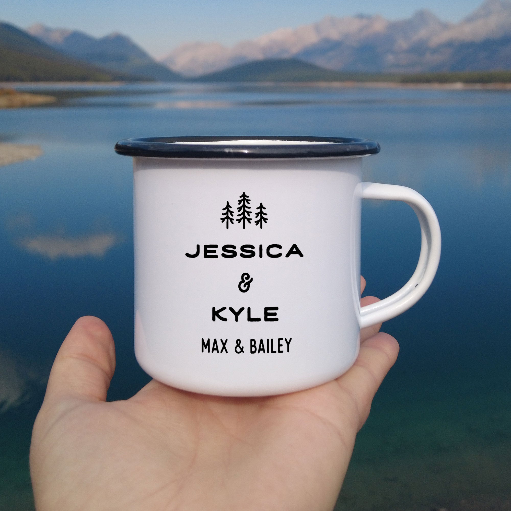 Close-up of hand holding single black rimmed enamel mug. Photo references the back (or right side) of the enamel mug design. The back has 3 Line Art Trees and the text 'Jessica & Kyle, Max & Bailey.'