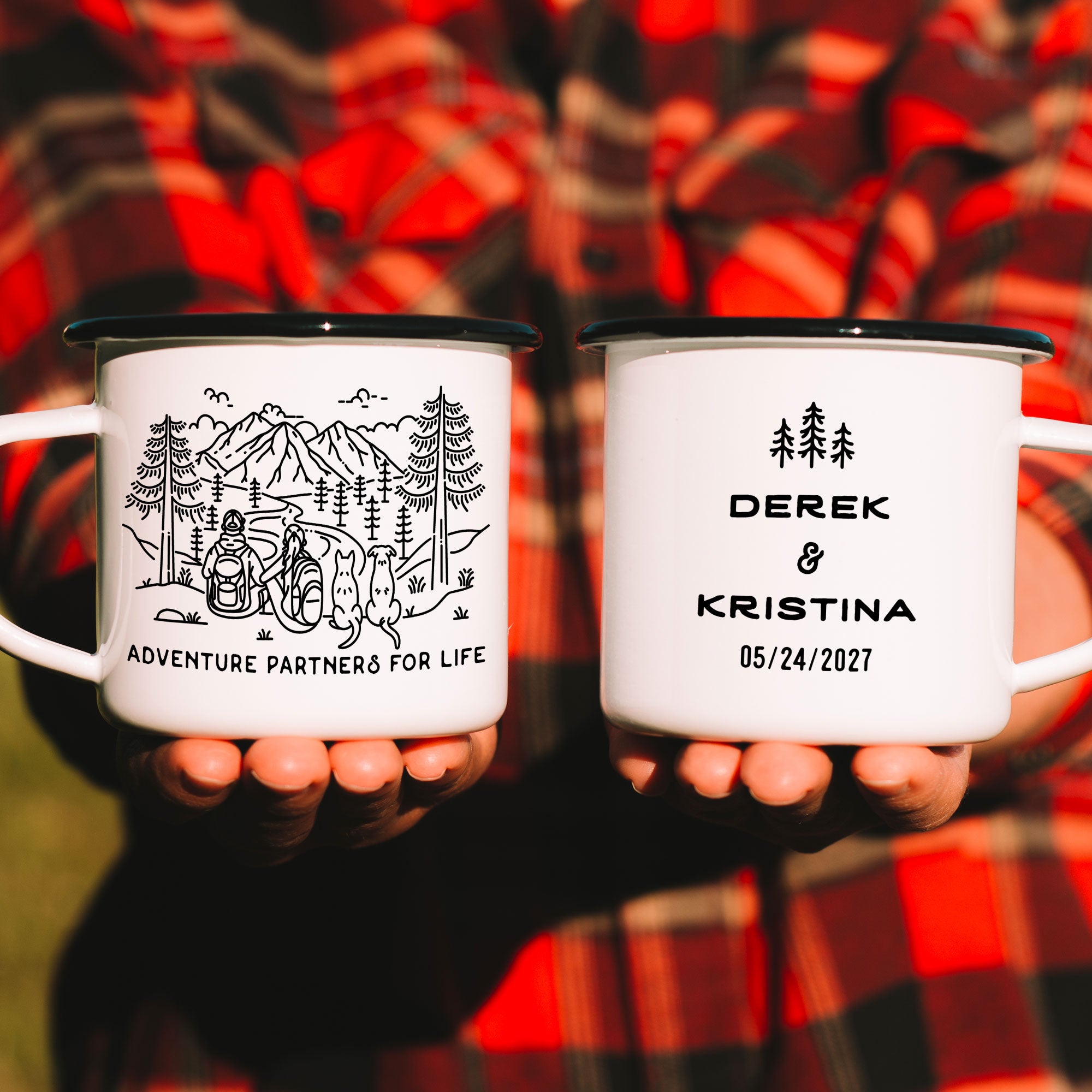 Hands holding two black rimmed enamel mugs. Features original hand drawn line artwork of mountain hiking couple & two dogs (1 pointy ear & 1 floppy eared). Text on front of mug shows 'Adventure Partners for Life'. Design on back of mug is of three line art trees and the text 'Derek & Kristina 05/24/2027.'