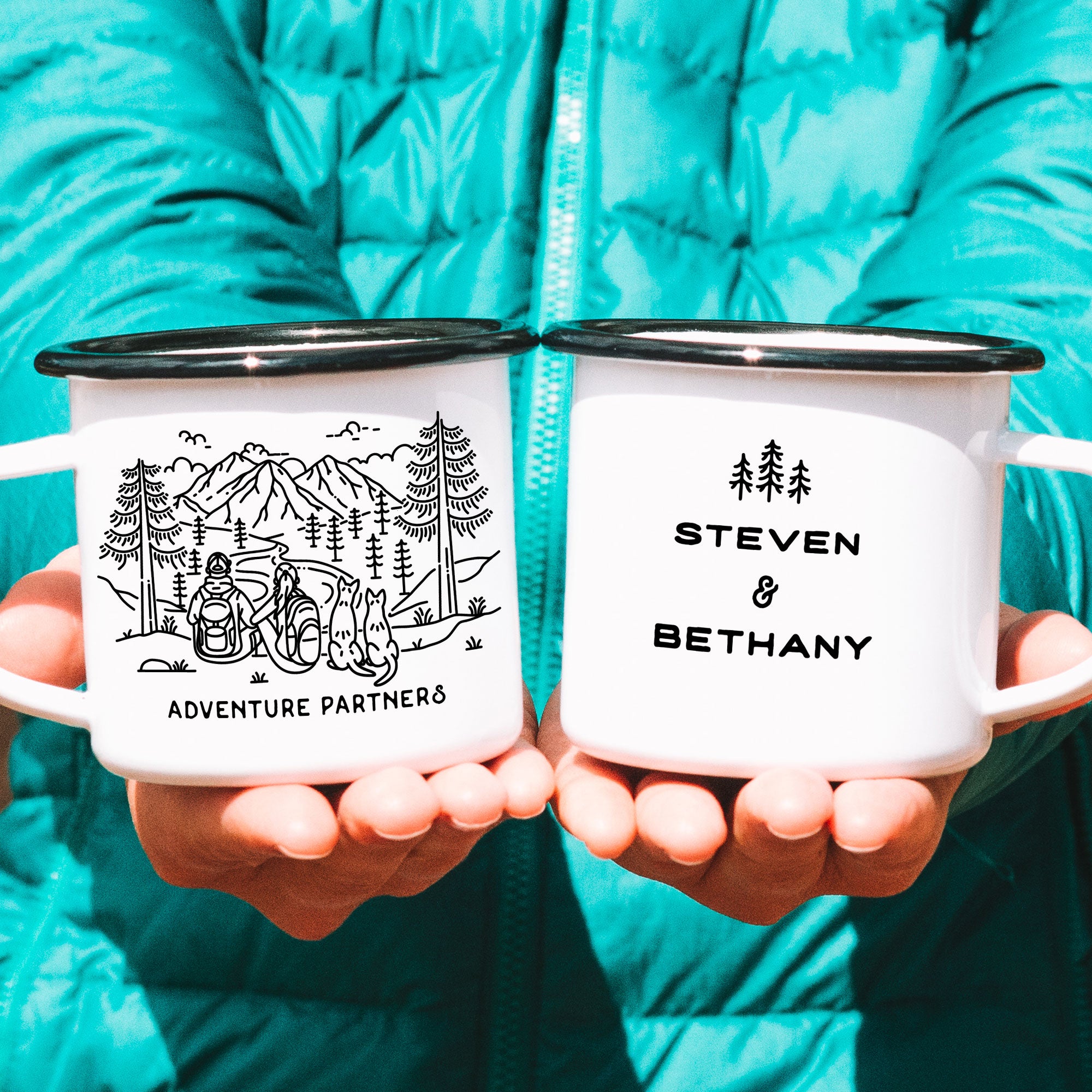 Hands holding two black rimmed enamel mugs. Features original hand drawn line artwork of mountain hiking couple & two pointy eared dogs. Text on front of mug shows 'Adventure Partners'. Design on back of mug is of three line art trees and the text 'Steven & Bethany'