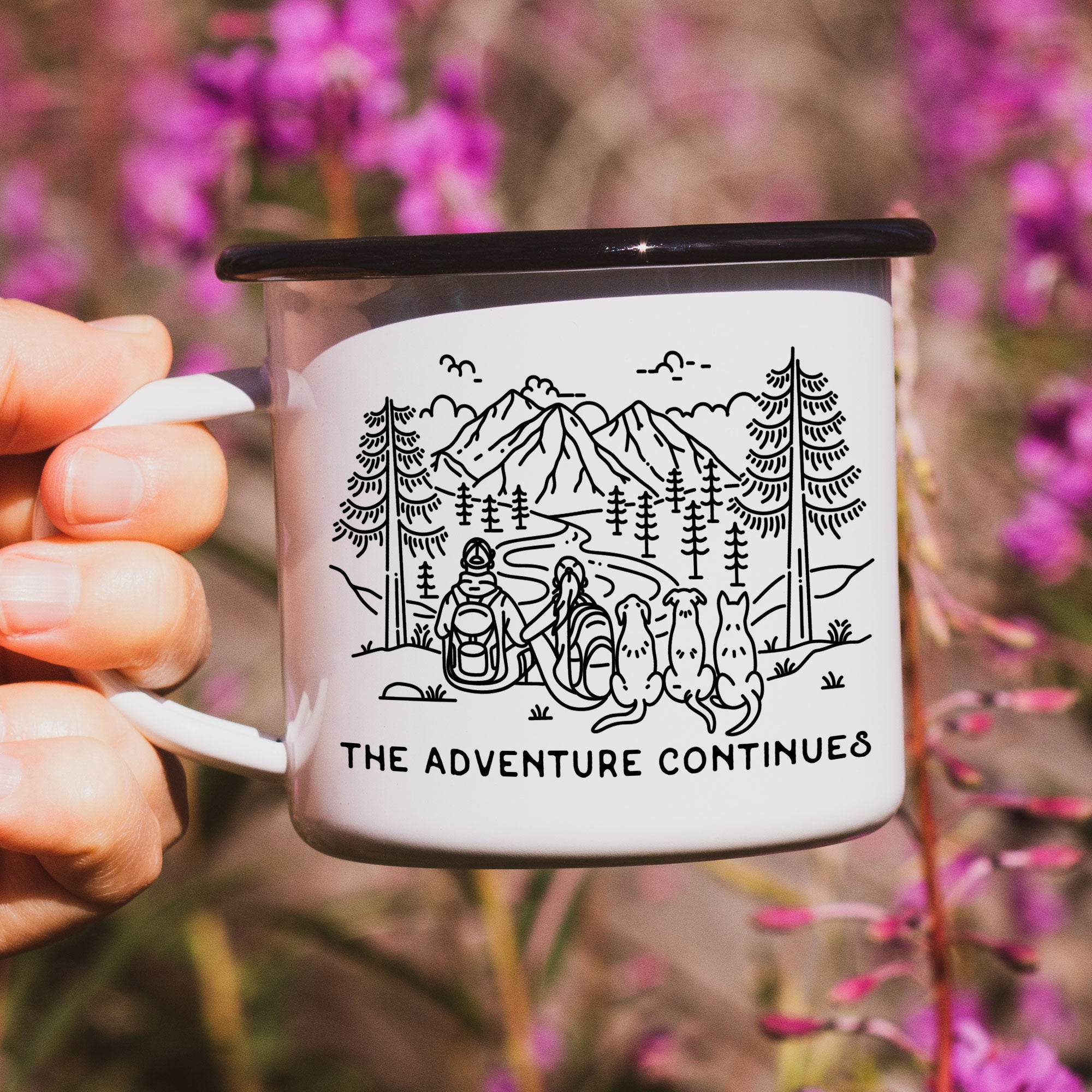 Close-up of hand holding single black rimmed enamel mug. The mug features line artwork of a mountain hiking couple with their 3 dogs. The front text reads The Adventure Continues.