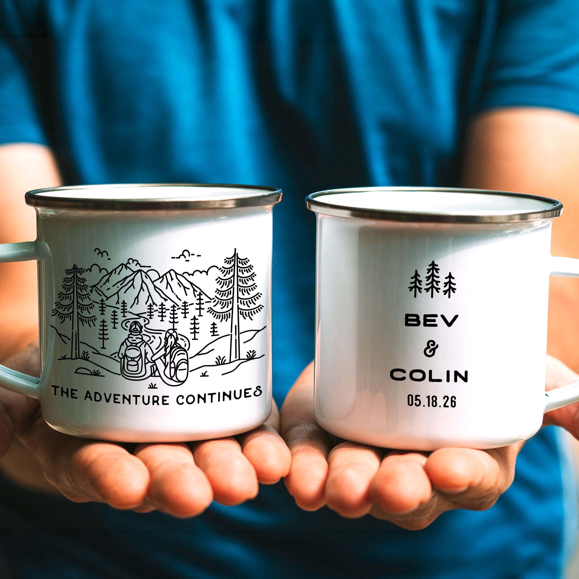 Man in blue shirt holding two silver rim enamel mugs. Desing of hiking man & woman looking out over mountains. Text reads underneath The Adventure Continues. Text on other mug (right side of mug) shows 3 Line Art Trees and text Bev & Colin 05.18.26