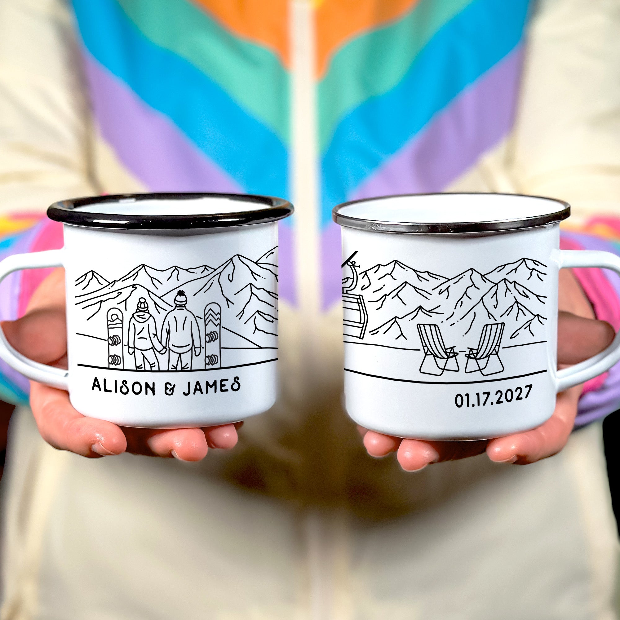 Hands holiding black rim & silver rim enamel mugs. Black rim mug shows snowboarding couple looking at mountain range. Text Alison & James on the bottom. Silver rim mug shows two ski chairs facing the mountains with the text 01.17.2027 on the bottom.