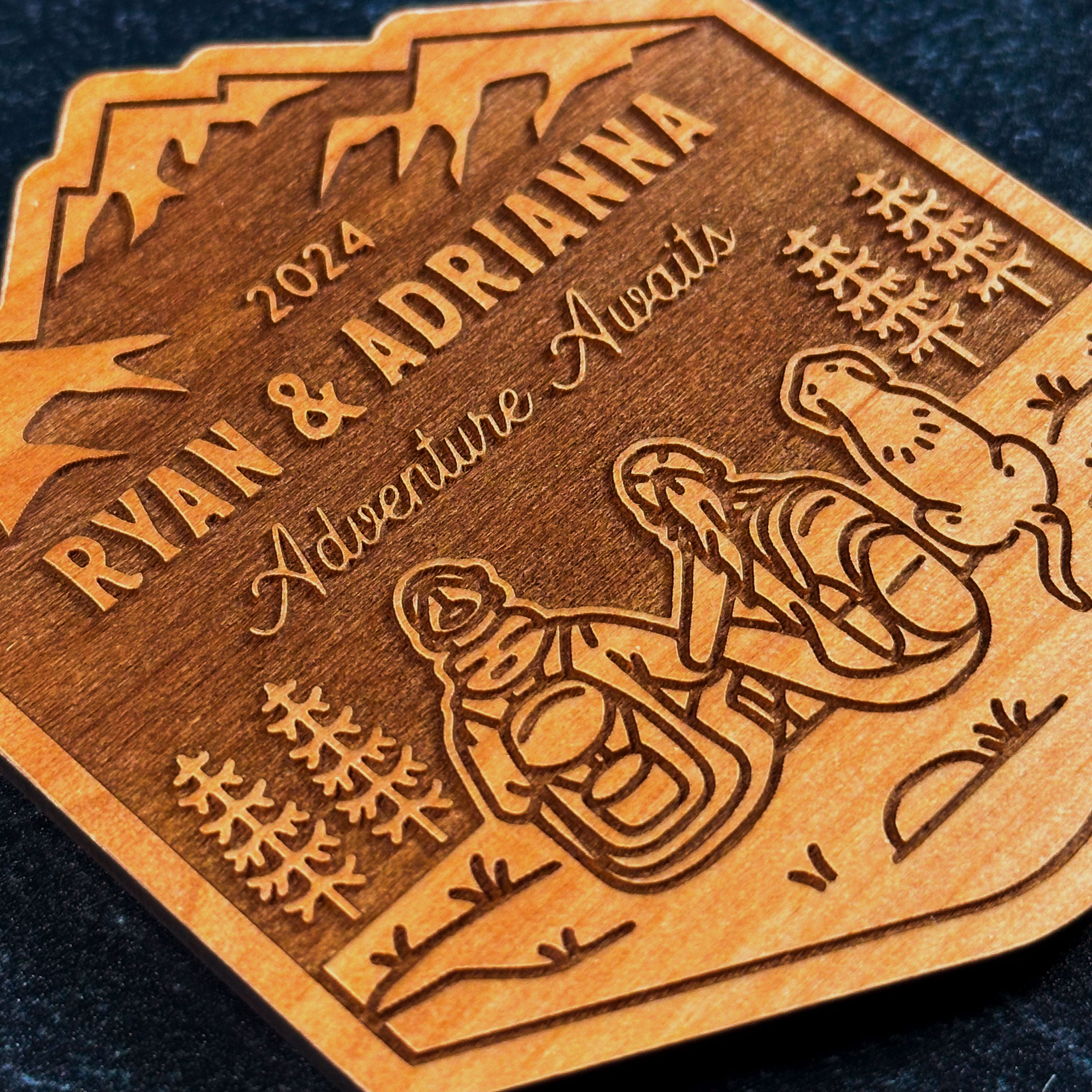 Engraved Wood Magnet closeup. Line artwork of hiking couple (man & woman) and floppy eared dog. Text reads 2024 (top), Ryan & Adrianna (middle), and Adventure Awaits in cursive (bottom)