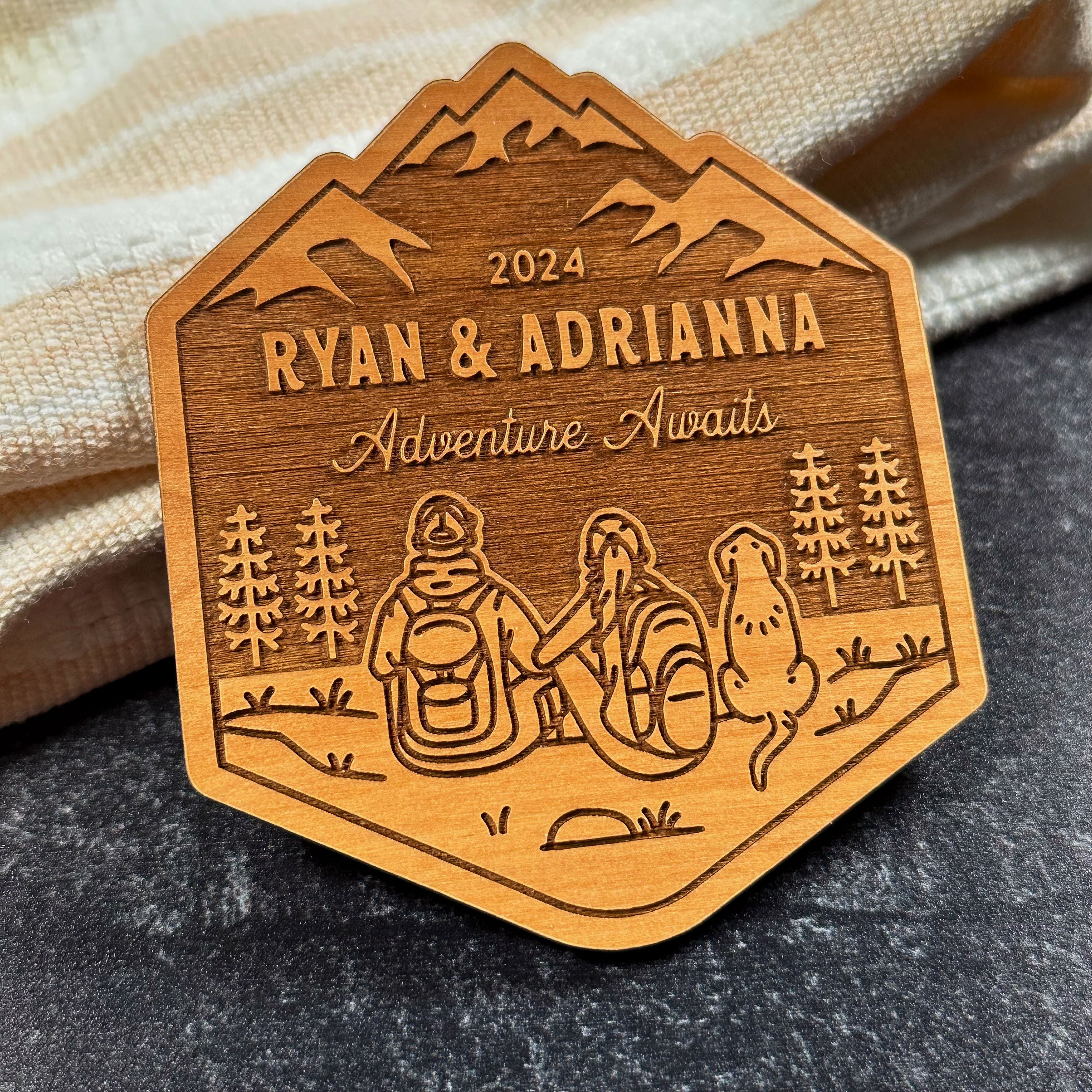 Wood magnet shaped like mountain. Line artwork of hiking couple (man & woman) and floppy eared dog. Text reads 2024 (top), Ryan & Adrianna (middle), and Adventure Awaits in cursive (bottom)